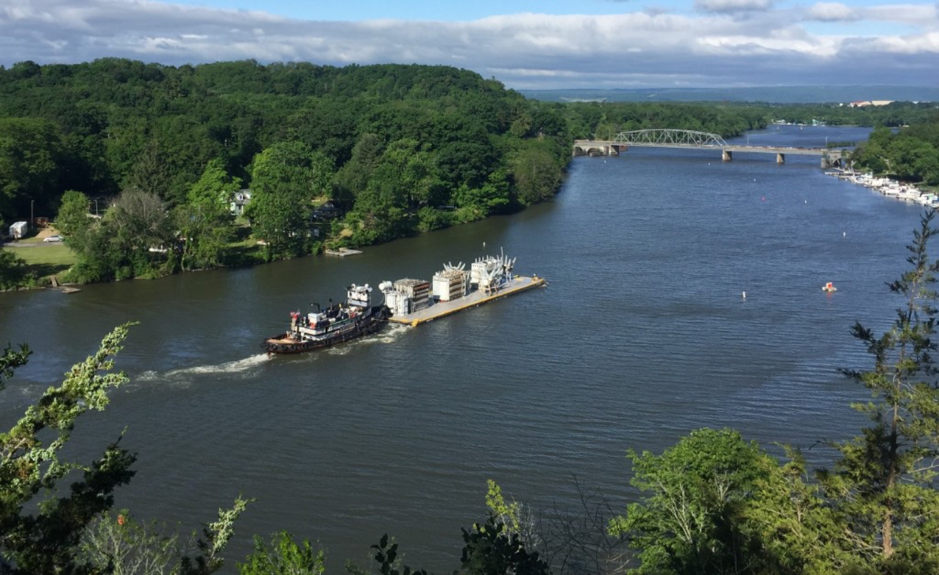 Tug pushing a deck barge with heavy electrical equipment on the Erie Canal, bridge and tree-lined shore in the distance.